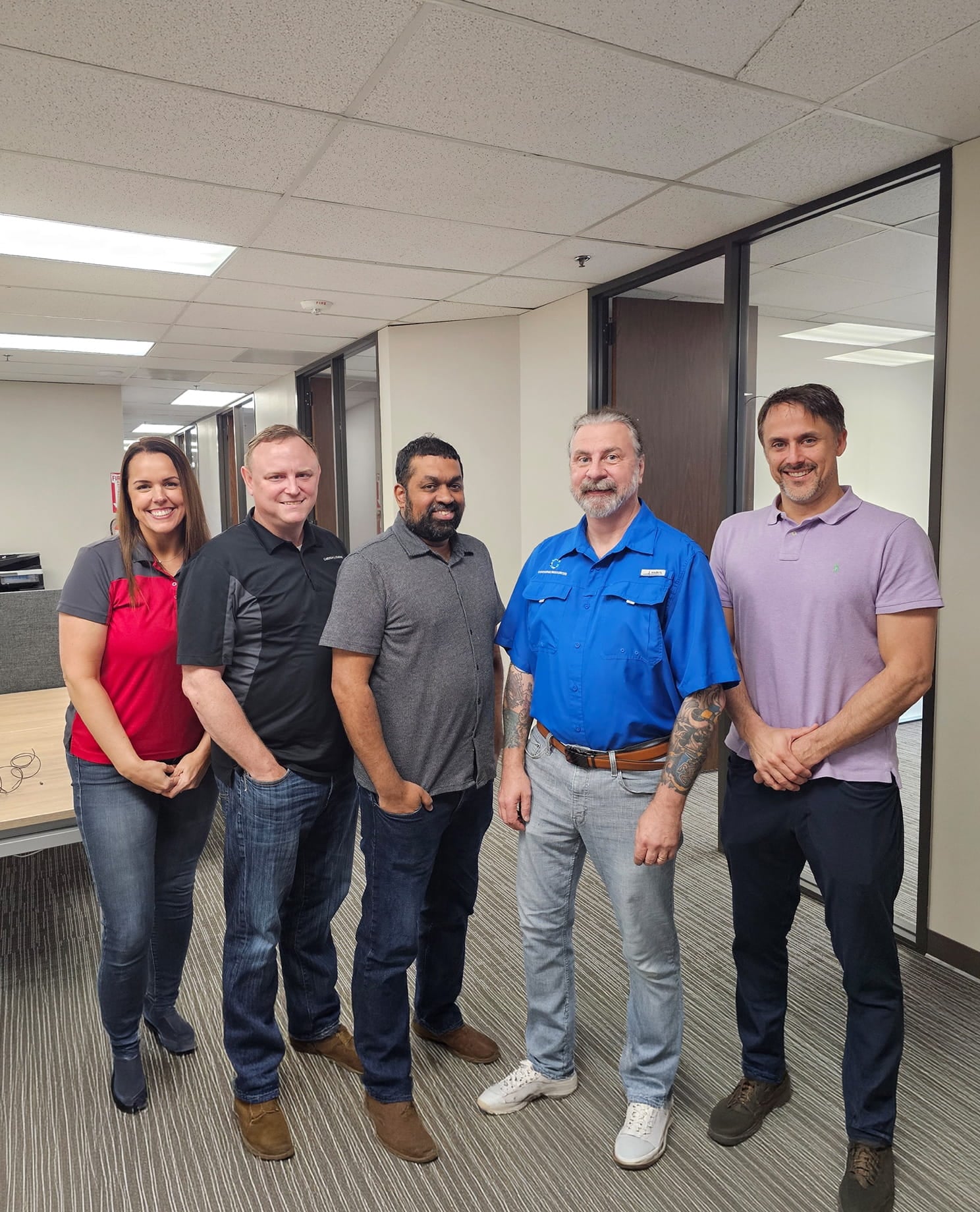 Six office colleagues, five men and one woman, stand smiling in a modern hallway.