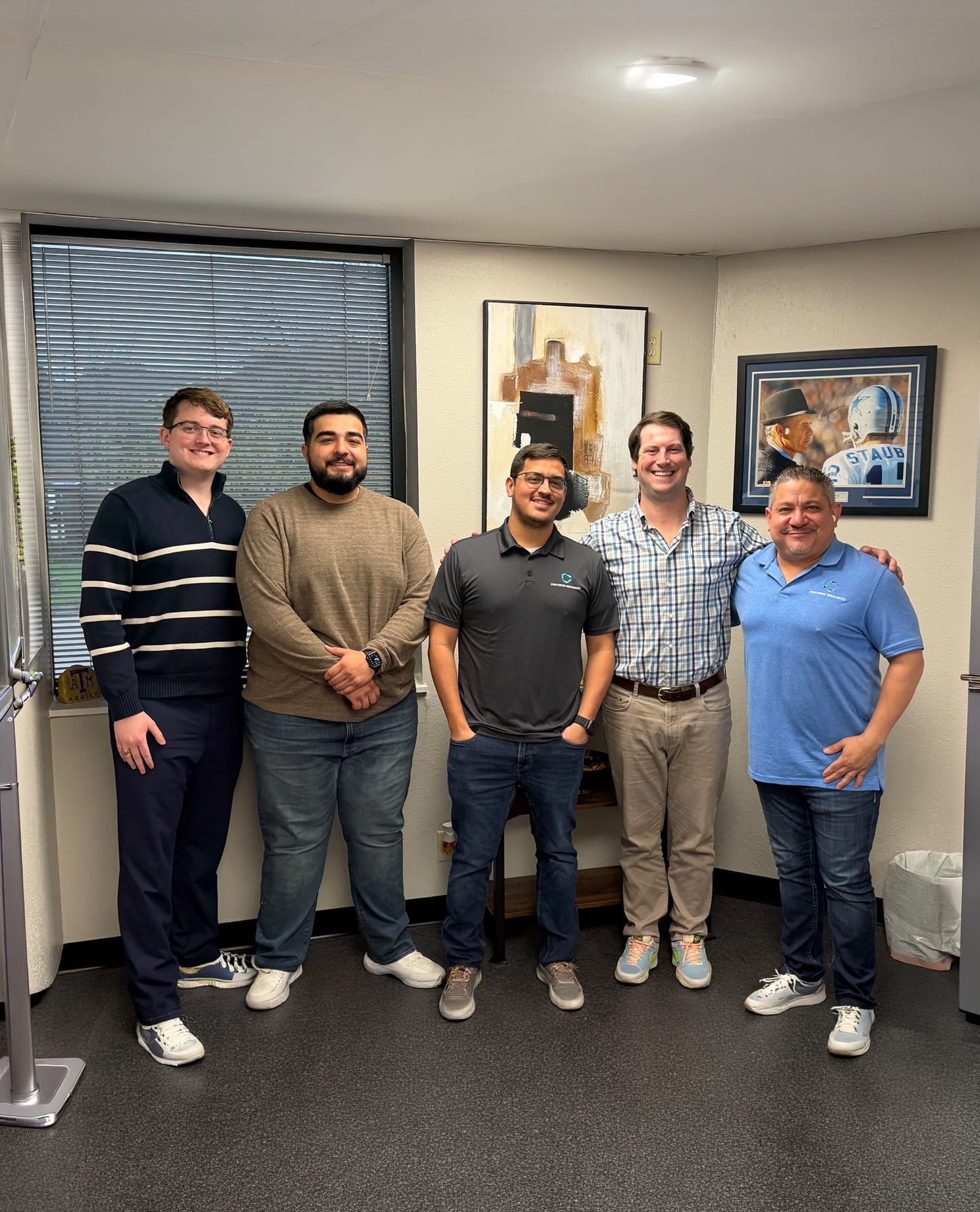 Six men posing together in an office, smiling, with a window, artwork, and a framed football photo on the wall.