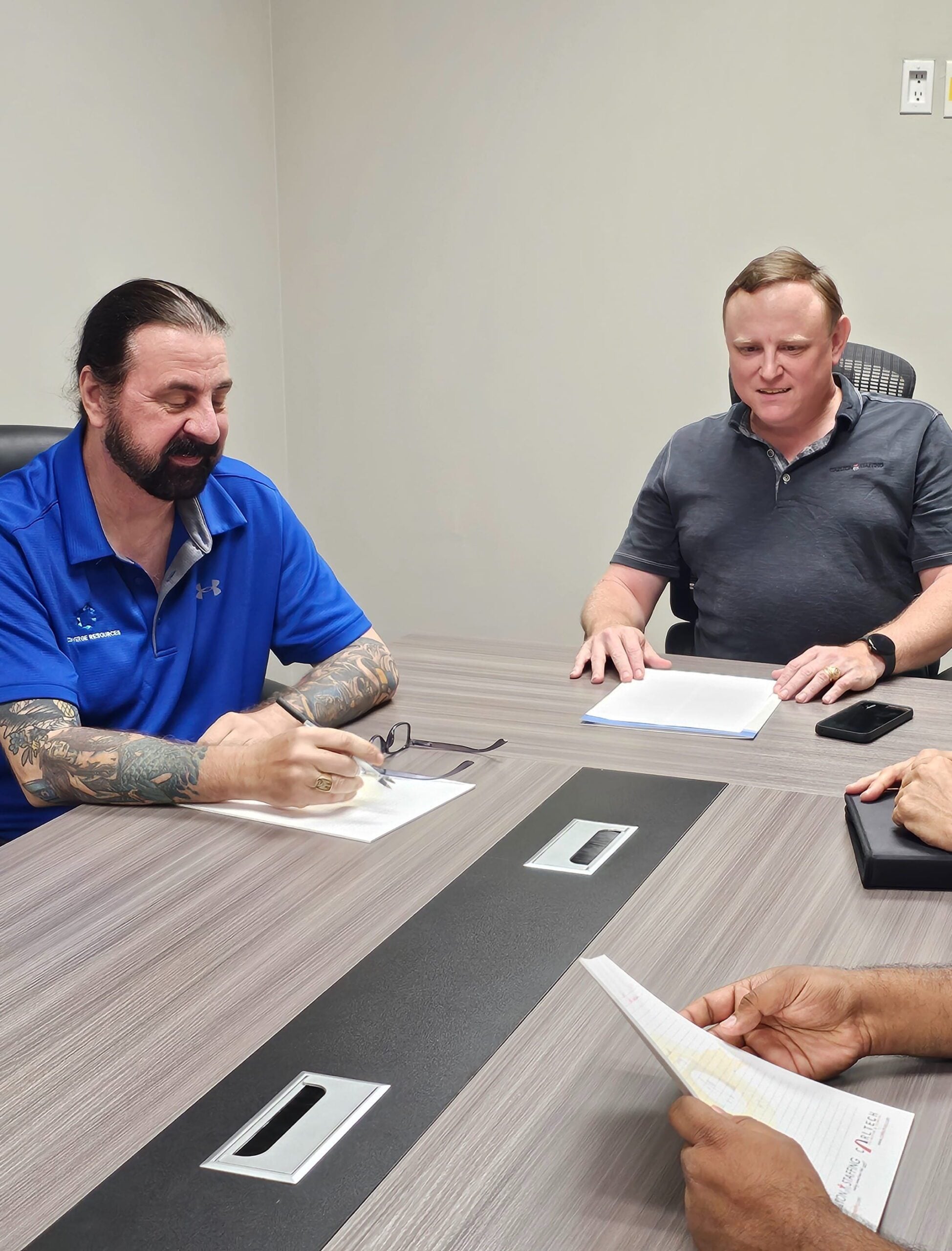 Two men at a meeting table; one in blue polo writes on paper, another in gray polo watches, third person holds a notepad.
