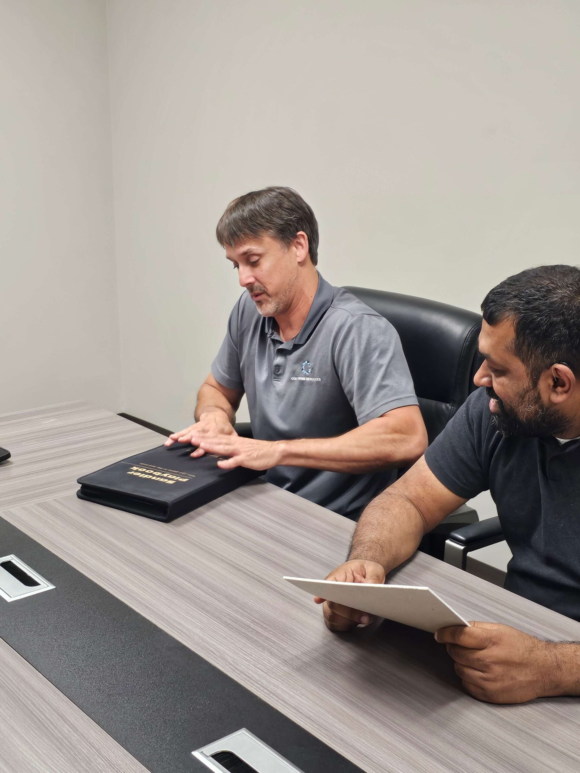 Two men at a conference table; one in a gray polo displays a black Sandler Playbook booklet, the other holds a white card.