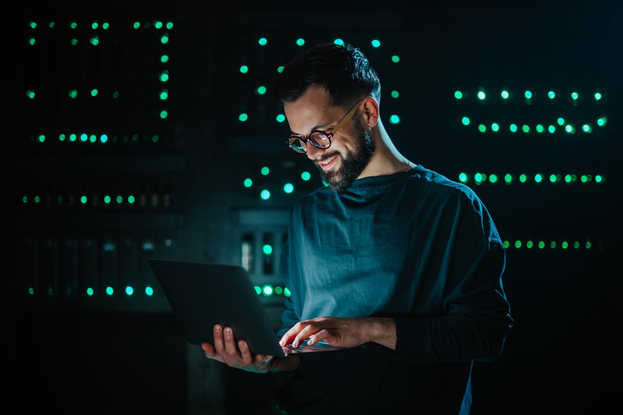 Bearded man with glasses smiles while using a laptop in a dim server room with green LED lights