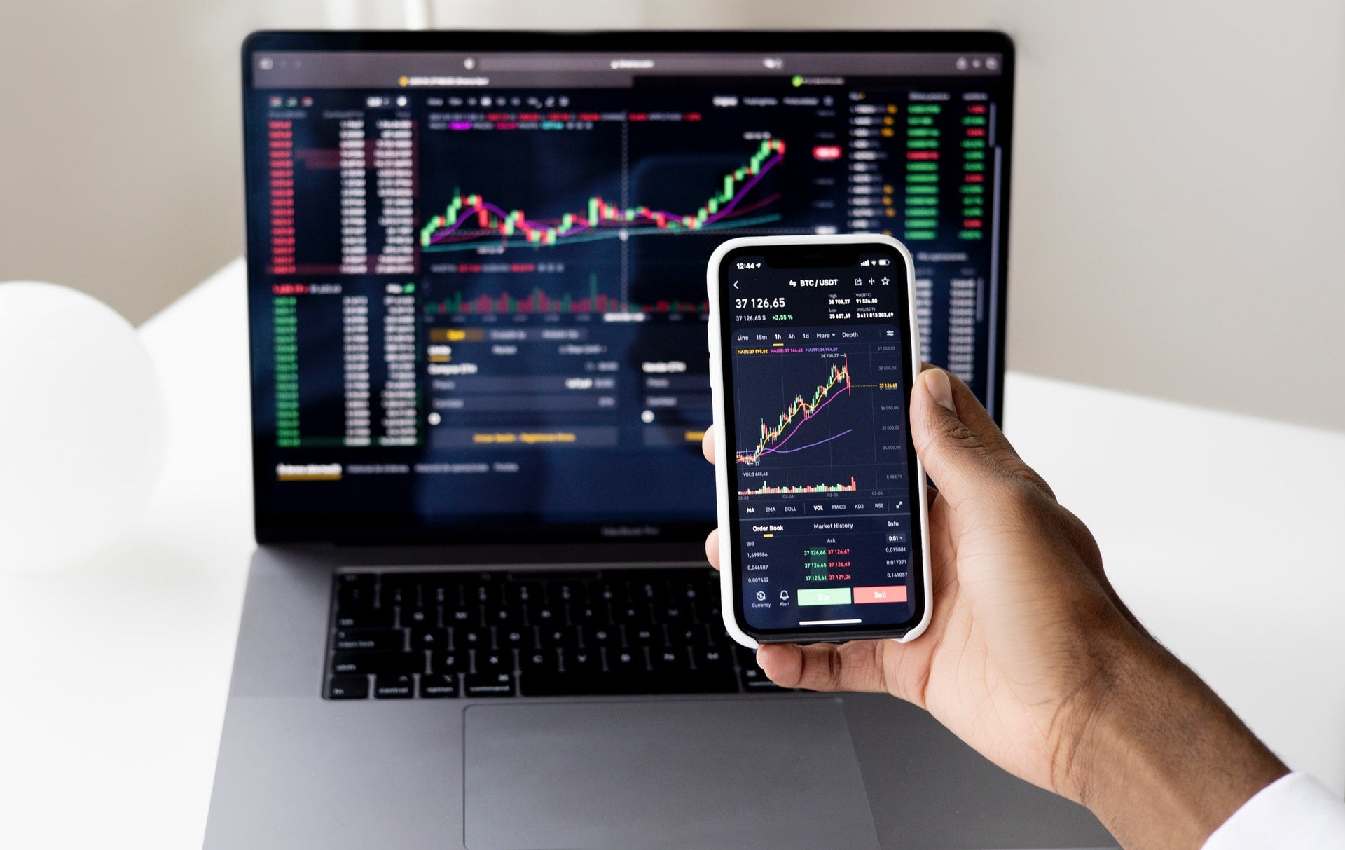 Hand holding smartphone displaying Bitcoin price chart, with laptop showing detailed trading interface in background.