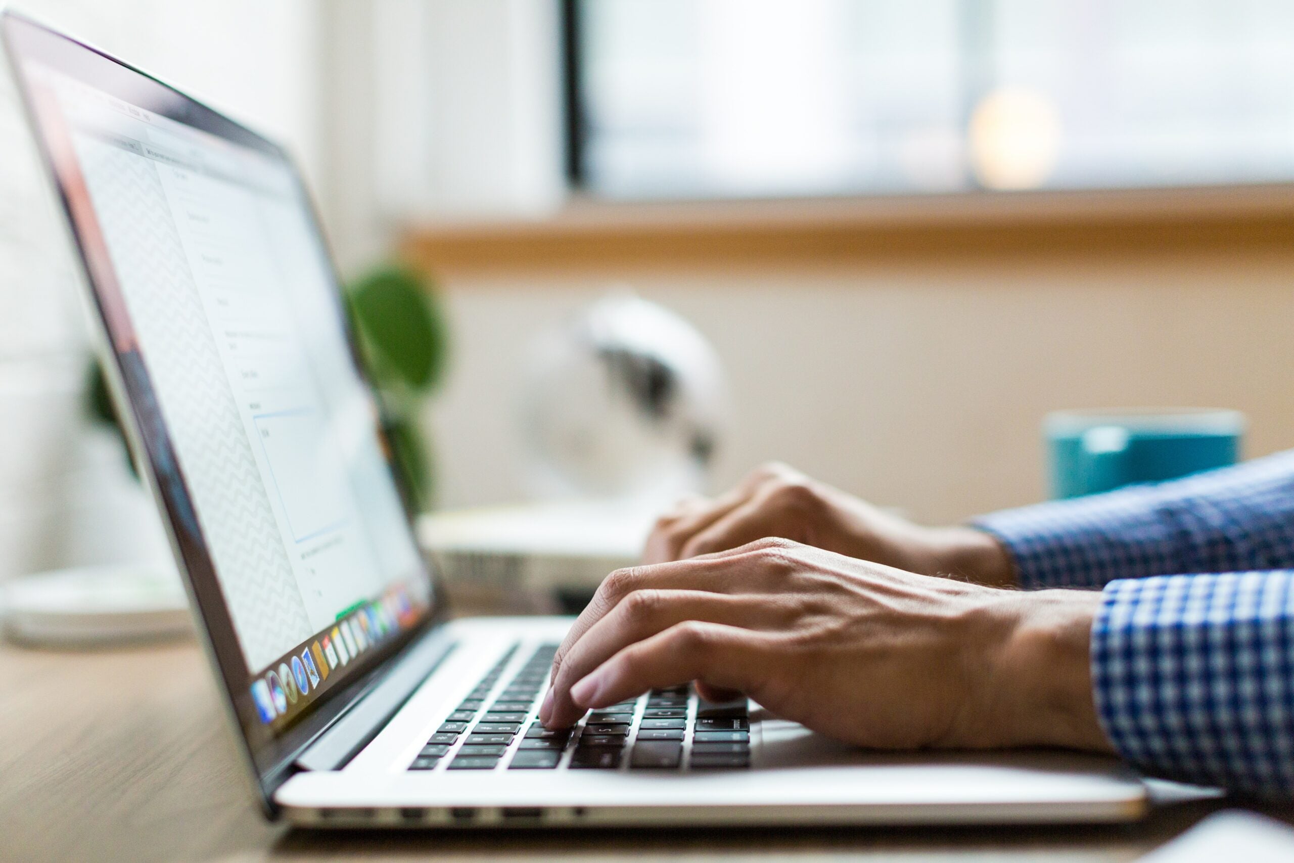 Person in blue checkered shirt typing on a silver laptop on a desk, blurred office background, window, plant, blue cup.