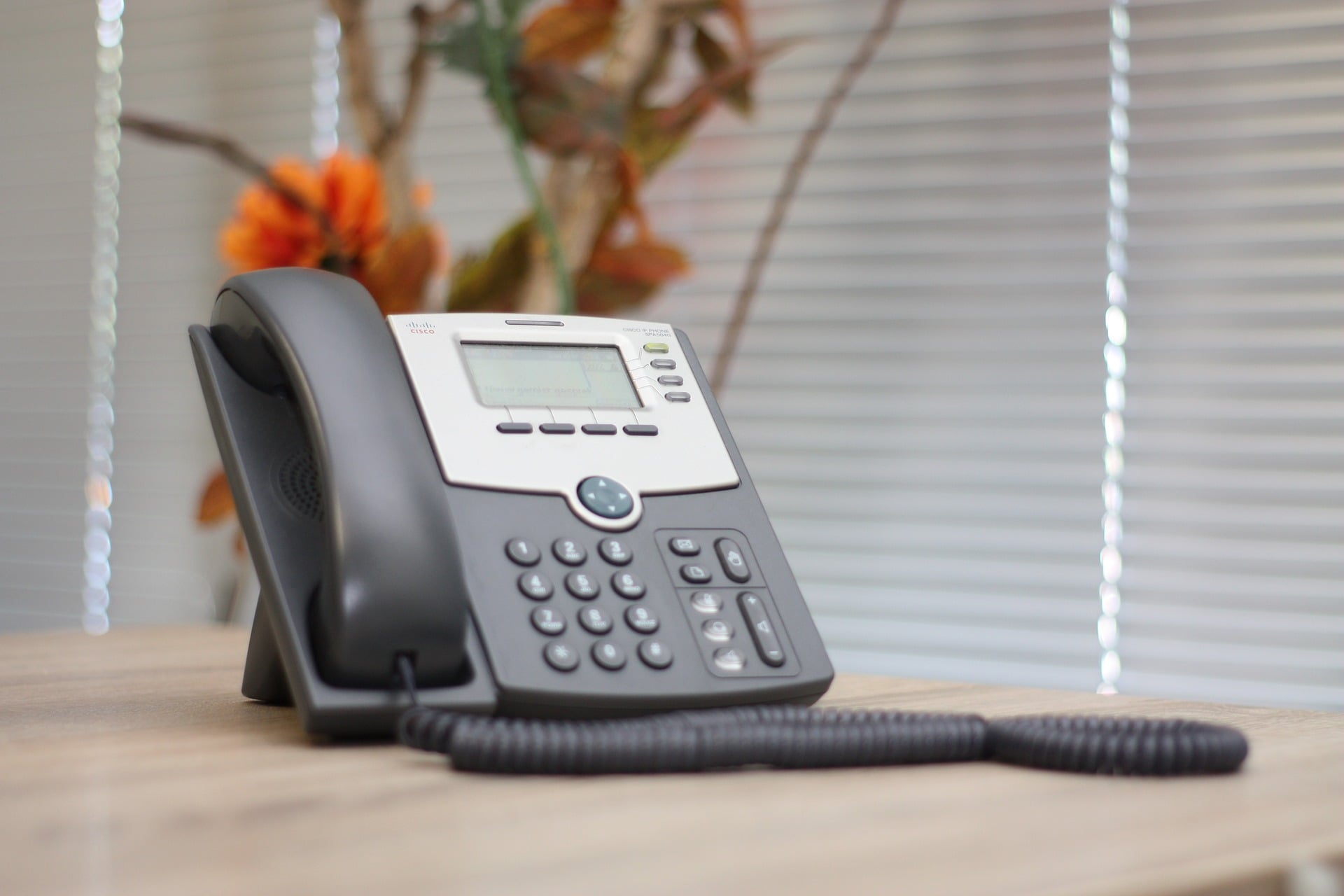 Cisco office phone with handset on wooden desk, digital display, keypad, set before window blinds and flowers.