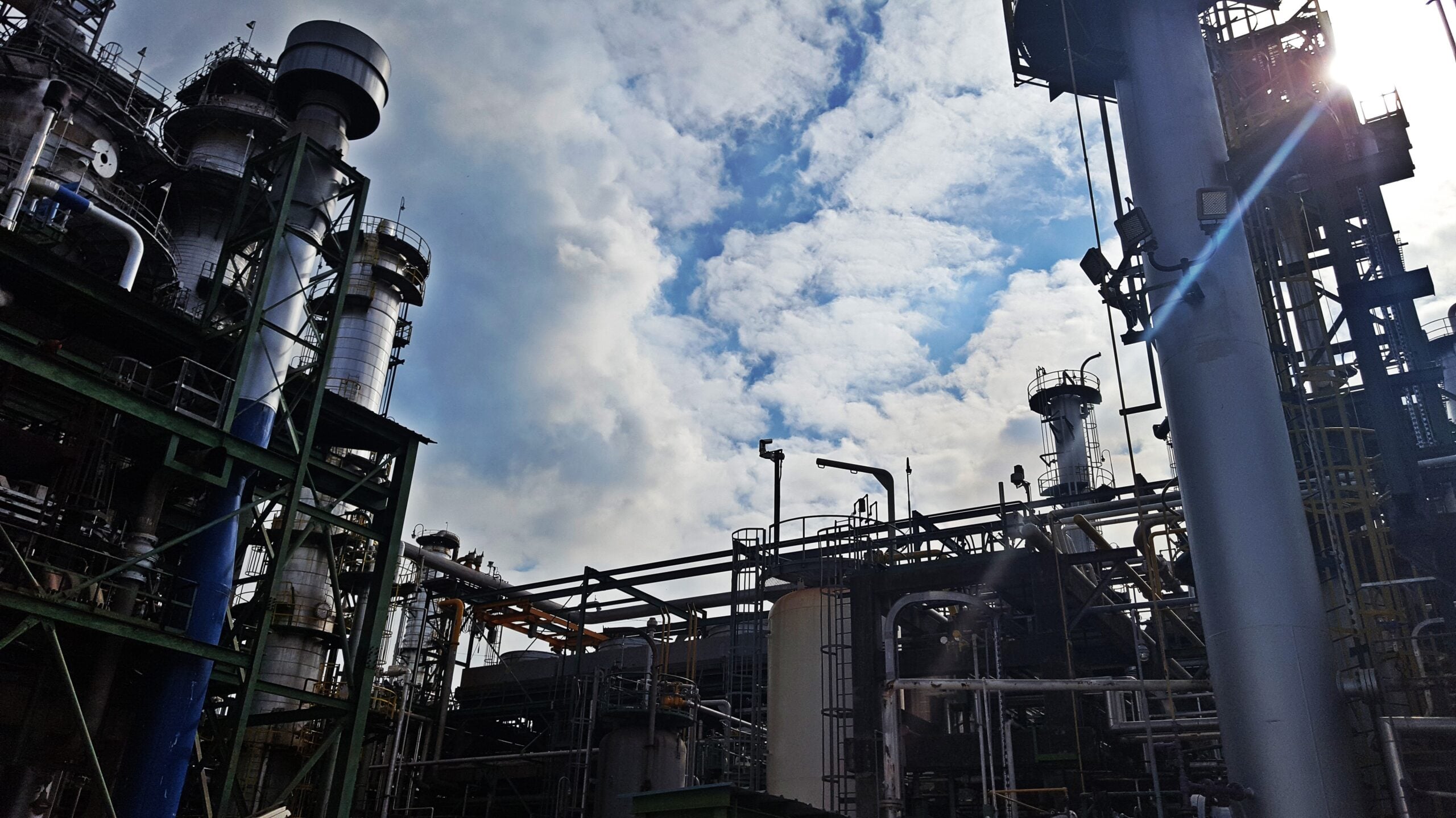 Industrial refinery complex with towering metal structures, pipes, and storage tanks under a cloudy blue sky.
