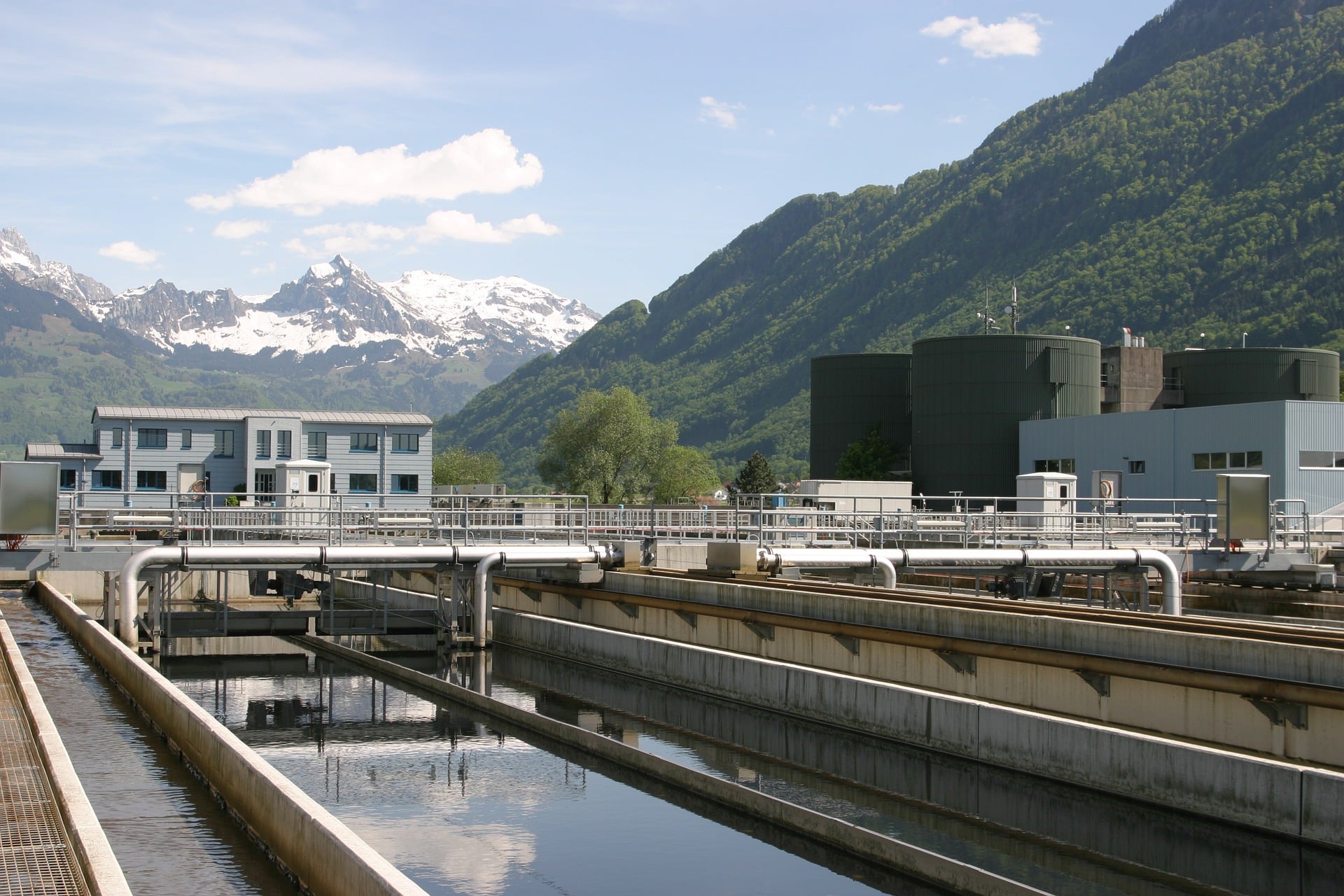 Water treatment plant with concrete channels, metal pipes, blue building, green hills, snow-capped mountains under blue sky.